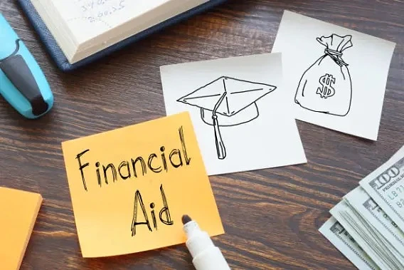 Dark wood table top with various objects. Partial view of several $100 bills, blue highlighter, yellow sticky notepad, financial ledger; 2 white sticky notes with a money bag symbol and graduation cap; yellow sticky note with Financial Aid written and marker laid on top